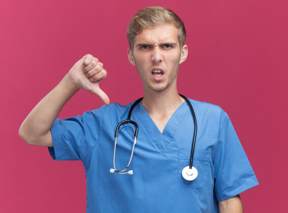 拇指年轻的男医生穿着医生制服 听诊器显示拇指朝下 隔离在粉红色的墙上年轻穿不愉快
