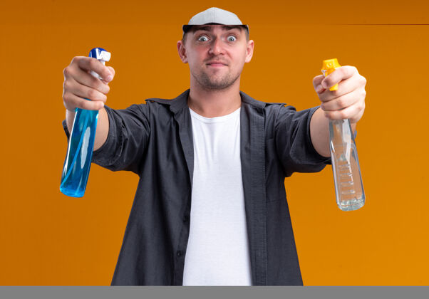 瓶子自信的年轻帅气的清洁工穿着t恤 戴着帽子 拿着喷雾瓶隔离在橙色的墙上帽子T恤清洁