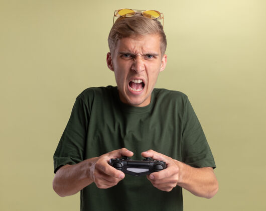 操纵杆愤怒的年轻帅哥身穿绿色衬衫 头戴眼镜 在橄榄绿的墙上玩游戏衬衫头眼镜