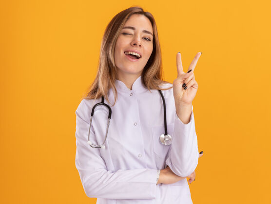 医疗阿米玲眨着眼睛 年轻的女医生穿着医用长袍 听诊器显示和平的姿态 隔离在黄色的墙上眨眼女性长袍