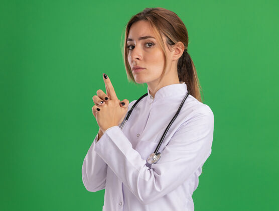 自信自信的年轻女医生穿着医用长袍 听诊器显示手势 隔离在绿色墙上展示医疗手势