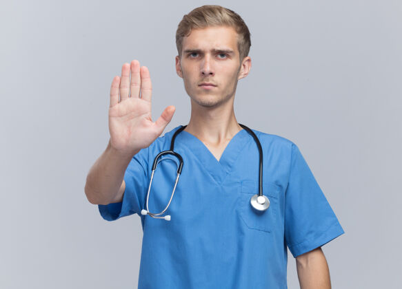 听诊器严格的年轻男性医生穿着医生制服 听诊器显示停止手势隔离在白墙上医生年轻男性