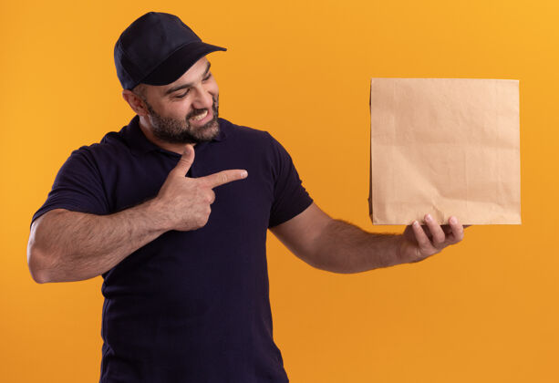 食物身着制服 戴着帽子 面带微笑的中年送货员指着隔离在黄色墙上的纸食品包装男人中年人点