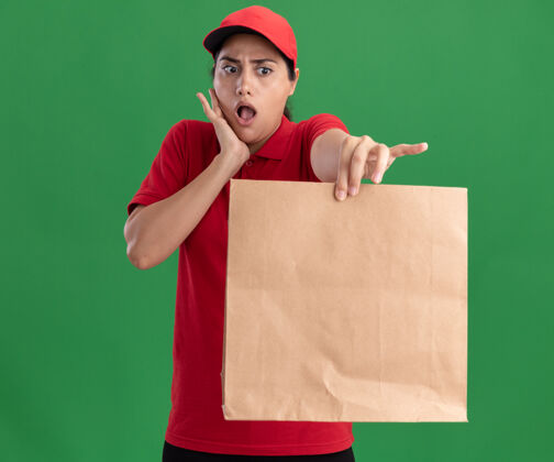 食品惊讶的年轻送货女孩穿着制服 戴着帽子 手里拿着纸食品包对着镜头 手放在隔离在绿色墙上的脸颊上举行纸张包装