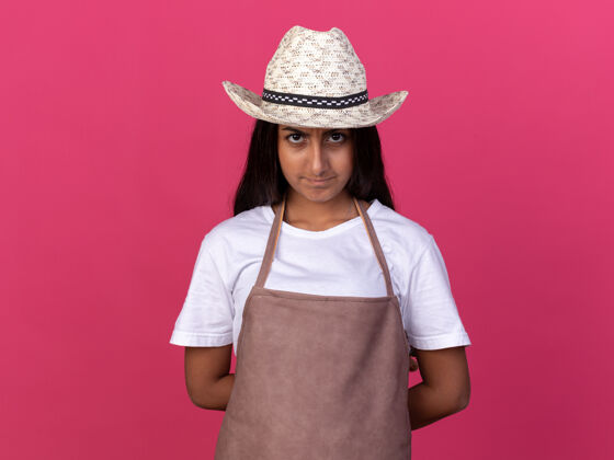 严肃的穿着围裙戴着夏帽的年轻园丁女孩 严肃的脸站在粉红色的墙上年轻的帽子围裙