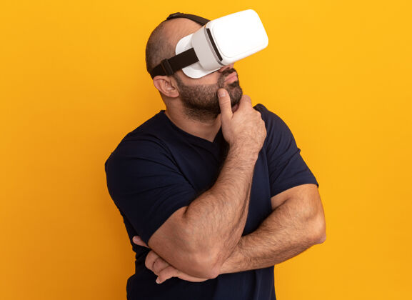 沉思留着胡子的男人穿着海军t恤 戴着虚拟现实的眼镜 手放在下巴上沉思着 站在橙色的墙上男人现实虚拟