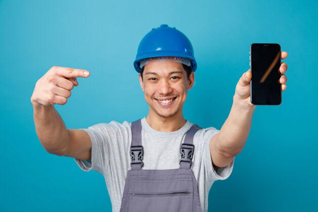 蓝色快乐的年轻建筑工人戴着安全帽 穿着制服 伸出手机对着摄像机指着它工人快乐朝向