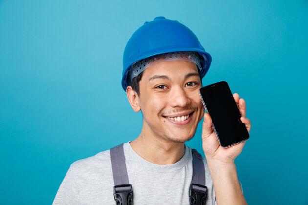 蓝色近距离观看微笑的年轻建筑工人戴着安全帽和制服手持手机观看电话安全