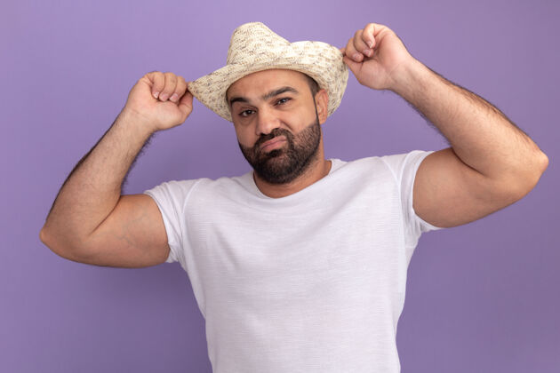 男人一位身穿白色t恤 头戴夏令帽 满脸胡须的男子站在紫色的墙上 用自信的表情抚摸着他的帽子帽子胡须立场