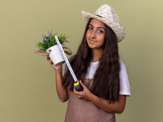 锅穿着围裙 戴着夏帽 拿着卷尺和盆栽植物 脸上带着微笑 站在绿墙上的年轻园丁女孩年轻微笑磁带