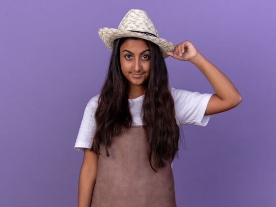 年轻穿着围裙 戴着夏帽 脸上带着自信微笑的年轻园丁女孩站在紫色的墙上帽子站着花园