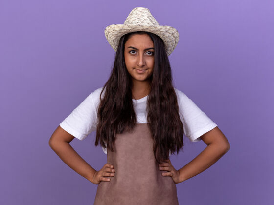 怀疑年轻的园丁女孩围着围裙 戴着夏帽 脸上带着怀疑的微笑站在紫色的墙上站着微笑围裙