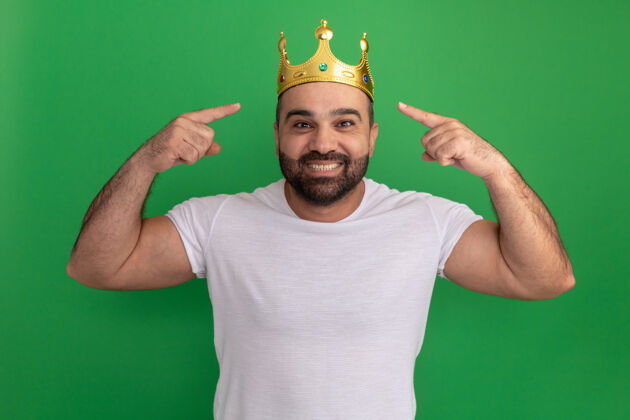 胡子戴着金色王冠的留胡子白t恤的男人开心地笑着用食指指着他站在绿色墙上的王冠目录手指皇冠