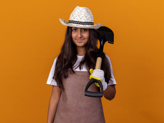 微笑年轻的园丁女孩拿着铲子微笑着站在橙色的墙上铲子站起来花园
