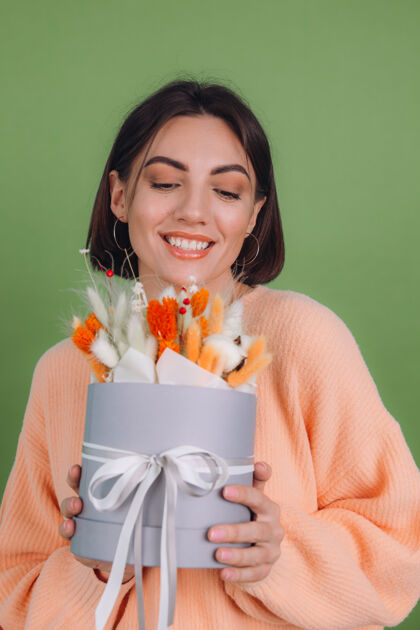 圆形年轻女子穿着休闲桃色毛衣隔离在绿橄榄墙上手持橙白色花盒组成的棉花 吉普赛拉小麦和拉古鲁斯作为礼物开心惊喜欢乐圆形盒静物