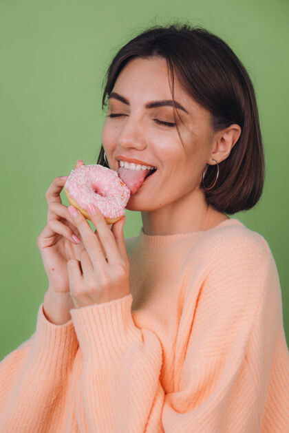 墙年轻时尚的女人穿着休闲的桃色毛衣孤立地站在绿橄榄色的墙上舔着粉色的甜甜圈复制空间女孩欢呼女人