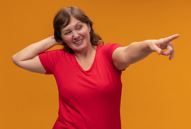 旁边一位身穿红色t恤的中年妇女 手放在头上 用食指指着站在橙色墙上微笑着的东西女人指向某物