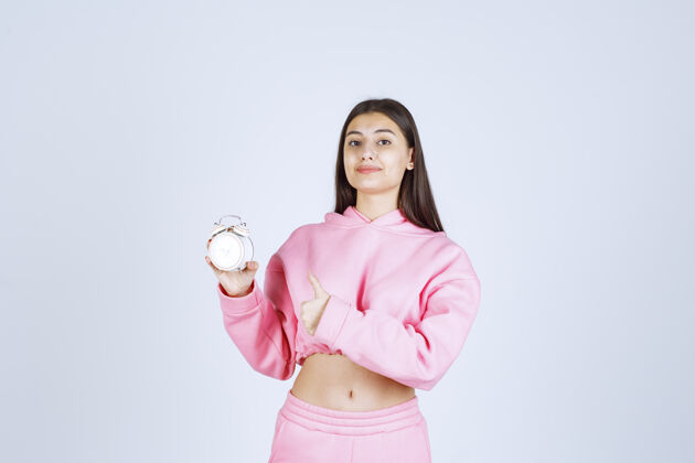 积极一个穿着粉色睡衣的女孩拿着一个闹钟 把它当作一个产品来推销休闲服装成人