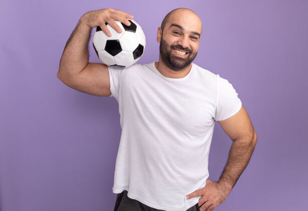 肩膀快乐的胡子男人穿着白色t恤 肩上扛着足球 微笑着站在紫色的墙上足球男人球