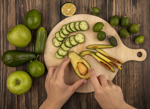 素食俯视图：女性用刀在木制菜板上切鳄梨 切碎的黄瓜片和整个黄瓜酸橙绿苹果和鳄梨隔离在木制表面蔬菜健康女性