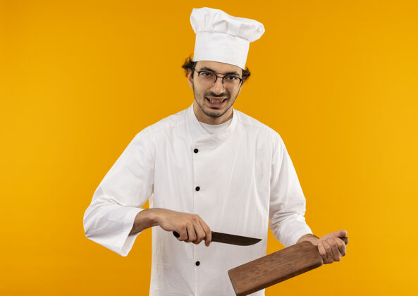 信心自信的年轻男厨师穿着厨师制服 戴着眼镜 手里拿着砧板和刀持有年轻男