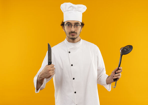 穿自信的年轻男厨师穿着厨师制服 戴着眼镜 手里拿着抹刀和刀自信黄色抹刀