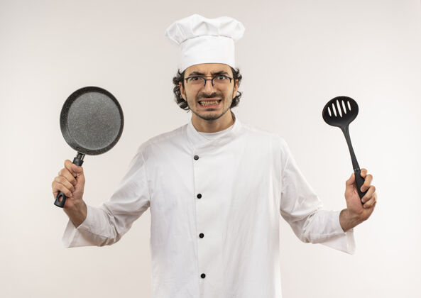 眼镜愤怒的年轻男厨师穿着厨师制服 戴着眼镜 手里拿着煎锅和抹刀制服穿男