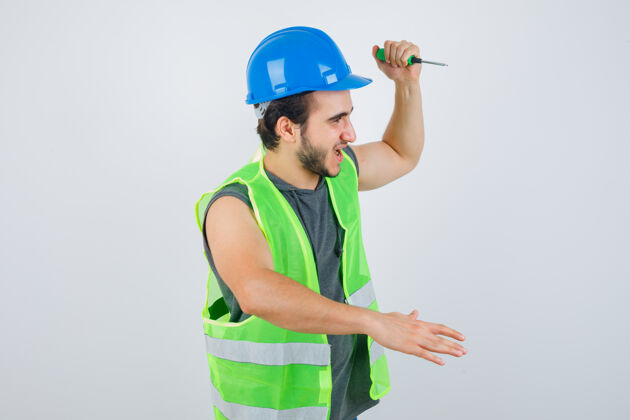 专业一幅年轻的建筑工人穿着制服举起手来用螺丝刀敲打的画像 看上去很疯狂不高兴英俊工人