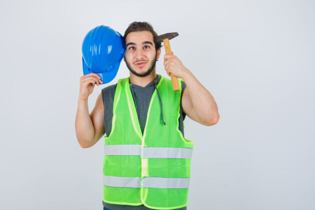 建筑商年轻的建筑工人拿着锤子和头盔 头上戴着工装制服 看上去很快乐 正对着前方学校工作服头盔