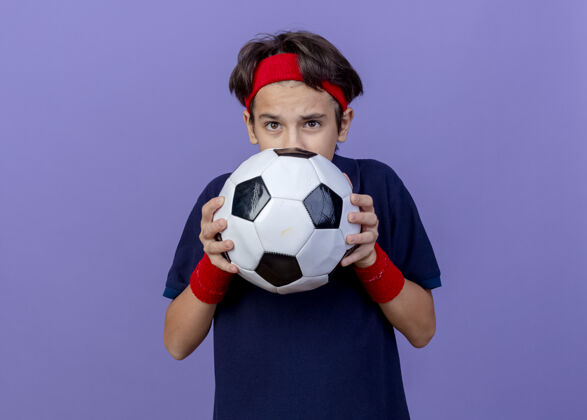 帅气令人印象深刻的年轻英俊的运动男孩戴着头带和护腕与牙套举行足球从后面它隔离在紫色墙上复制空间男孩戴运动