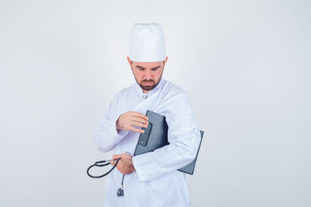 人年轻的男医生拿着剪贴板和听诊器 穿着白色制服 看上去很体贴 正前方视图交叉诊所听诊器