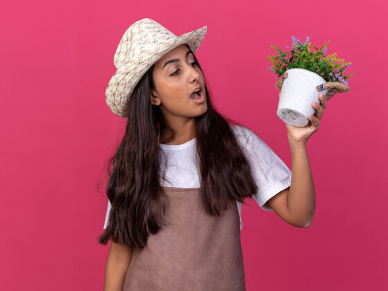 年轻穿着围裙 戴着夏帽 手里拿着盆栽植物的年轻园丁女孩站在粉色的墙上惊讶地看着它惊喜锅立场
