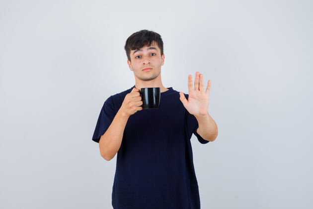 杯子一个穿着黑色t恤的年轻人手里拿着一杯茶 展示着停下来的姿势 看上去很害怕 正对着前方压力手手势