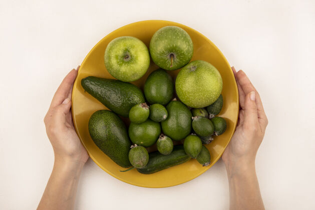 新鲜女性手拿着一盘黄色的新鲜水果的俯视图 比如苹果 菠萝和鳄梨 放在白色的表面上素食持有人