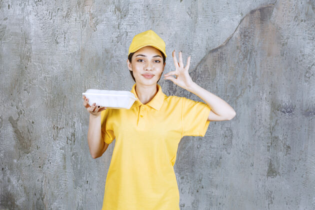 享受身着黄色制服的女服务人员手持一个塑料外卖盒 并显示积极的手势人体模特年轻交易