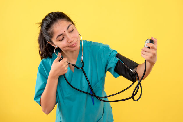 专业前视图漂亮的女医生穿着制服在黄色背景上使用血压测量仪使用听诊器医疗器械