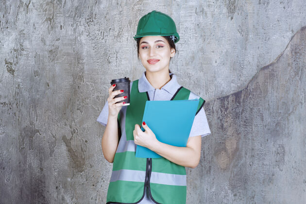 检查表身着绿色制服 头戴安全帽的女工程师 手里拿着一个黑咖啡杯和一个蓝色项目文件夹成人年轻人建筑师