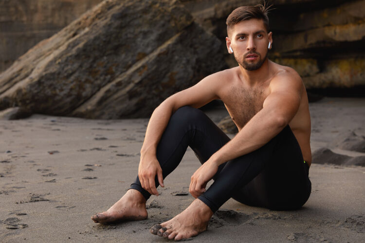 沙滩无忧无虑的男子冠军光着脚坐着 穿着黑色紧身裤 留着胡子射击音频赤脚