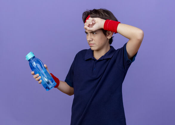 奶瓶疲惫的年轻英俊的运动男孩戴着头带和护腕 戴着牙套 手放在额头上 手举着 看着紫色墙上孤立的水瓶运动前额年轻
