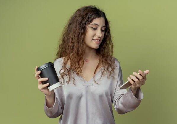 女性年轻漂亮的女上班族拿着一杯咖啡 看着橄榄绿墙上孤立着的手里的电话杯子手年轻