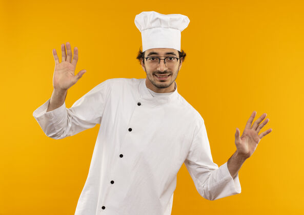 制服面带微笑的年轻男厨师身着厨师制服 戴着眼镜 双手孤立地摊开在黄色的墙上手烹饪传播