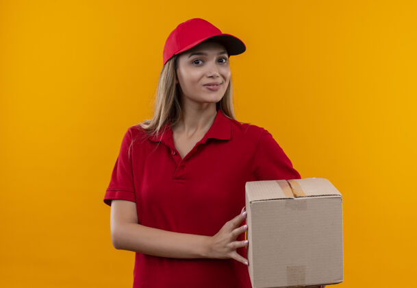 微笑微笑着的年轻送货女孩穿着红色制服 戴着一个装帽子的盒子 被隔离在橙色的墙上帽子戴着拿着