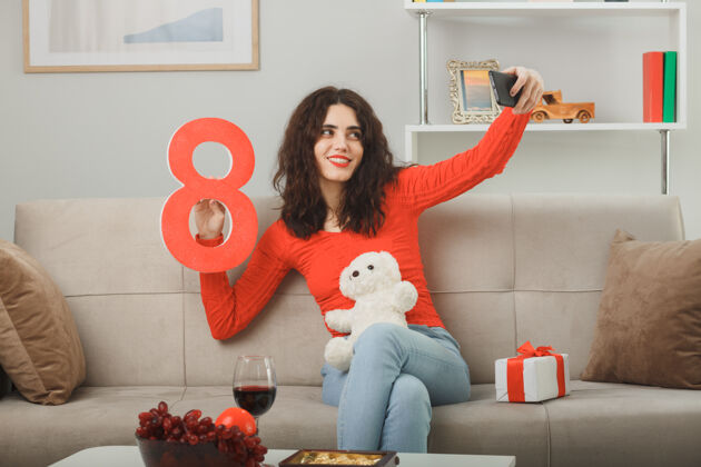 休闲穿着休闲服的快乐年轻女子坐在沙发上拿着8号礼物 泰迪熊在用智能手机自拍 微笑着庆祝三八国际妇女节庆祝自拍坐着