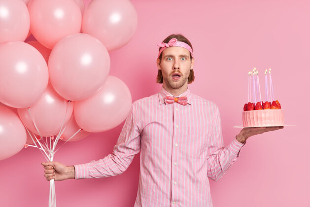 人惊喜的生日男子震惊地收到这么多来自亲朋好友的祝贺与气球和节日蛋糕摆姿势身着衬衫蝴蝶结头带孤立在粉红色的墙上乐趣祝贺帅哥
