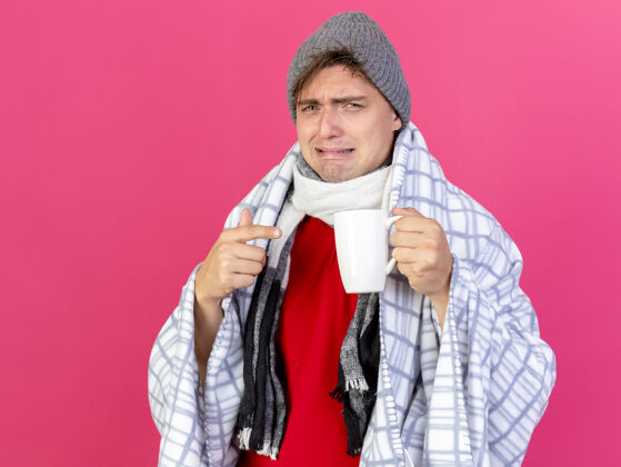 男人恼怒的年轻英俊的金发病男子戴着冬天的帽子和围巾包裹在格子布举行 并指着杯子隔离在深红色的墙壁与复制空间格子穿疾病