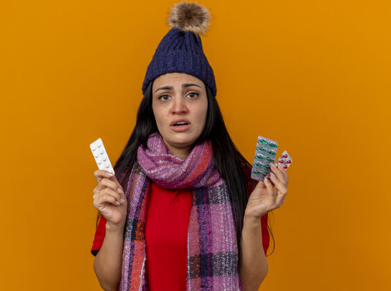 帽子年轻的高加索病女孩戴着冬天的帽子和围巾 手里拿着几包药片 隔离在橙色的墙上 留着复印空间药片围巾不愉快