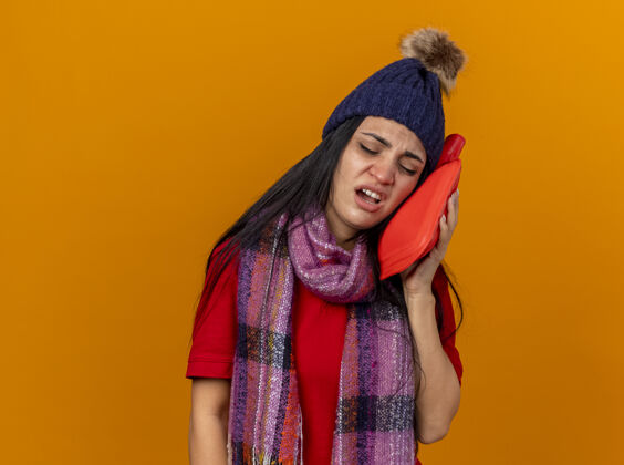热疼痛的年轻白种人生病的女孩戴着冬天的帽子和围巾用热水袋触摸脸闭着眼睛隔离在橙色的墙上有复印空间触摸橙色帽子