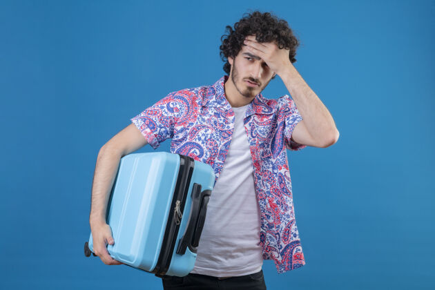 累了疲惫的年轻英俊的旅行家 手放在前额的手提箱上 放在与世隔绝的蓝色墙壁上 留有复制空间抱着年轻手提箱