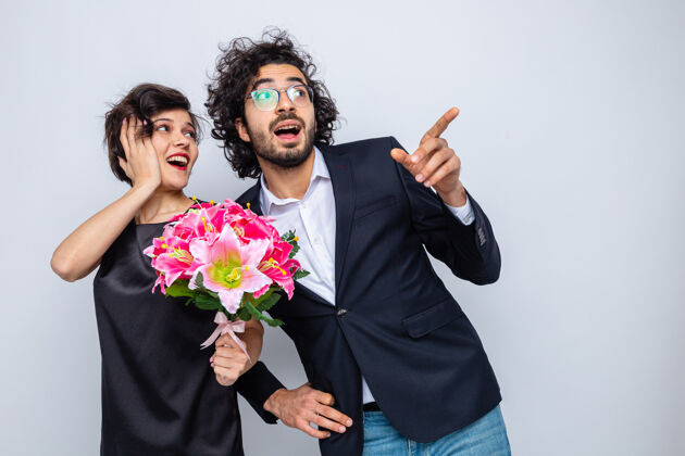 3月8日年轻漂亮的夫妇幸福的男人和女人拿着花束看着一边高兴和惊讶地用食指指着一边庆祝3月8日国际妇女节日国际男人