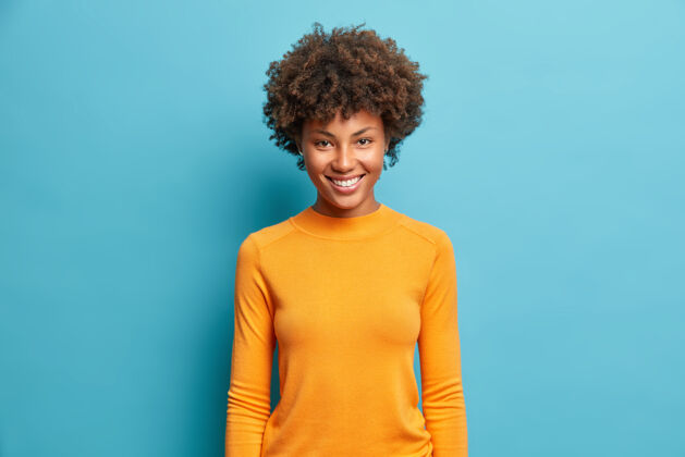 乐观半身镜头中 一位年轻而开朗的女士穿着休闲的橙色套头衫 脸上洋溢着愉快的笑容 站在蓝色的墙上有趣健康爱